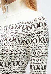 Adīts džemperis ar augstu apkakli, krēmkrāsas pamatu un sīkām brūnām ģeometriskām rakstām, tostarp viļņotām un rombu formām.