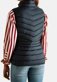 Czarna kamizelka puchowa z pikowaniem w wzór chevron, wysoki kołnierz i dopasowany fason, noszona na czerwonej i białej w paski koszuli. U dołu widoczne dżinsy.