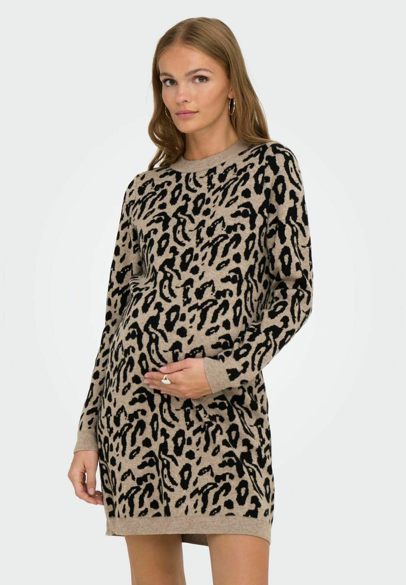 Femme enceinte portant une robe longue à manches longues beige et noire à motifs léopard, caressant doucement son ventre, se tenant devant un fond uni.