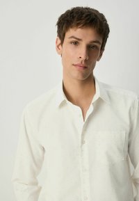 Cămașă albă cu guler clasic, având un model discret cu puncte, buzunar aplicat și textură netedă, purtată de un model cu păr scurt.