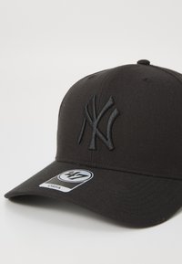 Svart baseballcaps med en hevet, svart brodert NY-logo. Har en flat skygge og en 47-merketang. Laget av stoff med en jevn tekstur.