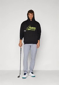 Čierna mikina s kapucňou s neónovo žltým grafickým textom, sivé nohavice a biele golfové topánky. Drží golfovú palicu, stojí na neutrálnej pozadí.