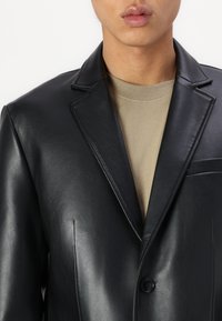 Schwarze Lederblazer mit Reverskragen, einfacher Knopfverschluss und subtiler Textur, getragen über einem beigen Rundhalsausschnitt-Shirt.