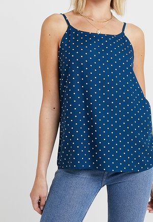 Haut sans manches bleu à pois avec fines bretelles, porté avec un jean bleu clair et des colliers dorés superposés.