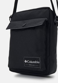 Czarna torebka typu crossbody z klapką, wyposażona w przedni guzik, naszywkę z logo i regulowany pasek. Kompaktowy i prostokątny kształt.
