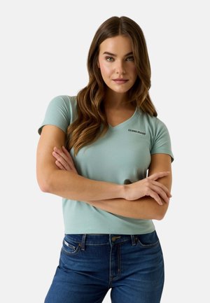 Giovane donna con lunghi capelli castani indossa una maglietta Guess Jeans azzurra a scollo a V e jeans blu scuro, con le braccia incrociate.