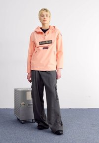 Jachetă impermeabilă roz deschis, cu glugă, buzunar frontal și detaliu cu logo. Pantaloni largi cu dungi negre și gri. Textură netedă, croială lejeră.