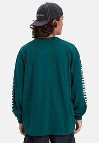 Chemise à manches longues en teal en coton, avec un col rond et un texte graphique blanc le long de la manche droite. Texture douce, coupe ample.