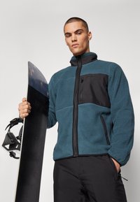 Chaqueta de forro polar azul con panel frontal negro, bolsillos con cremallera y cuello alto. El modelo sostiene una tabla de snowboard negra y fijaciones.