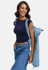 Lächelnde Frau mit welligem, dunklem Haar, die ein marineblaues Crop-Top und blaue Jeans trägt und eine hellblaue Jacke über ihrer Schulter trägt.