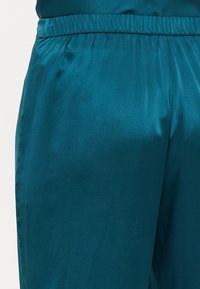 Teal satengbukser har et elastisk liv og en glatt, skinnende tekstur. Designet er løstsittende, med en subtil glans over stoffet.