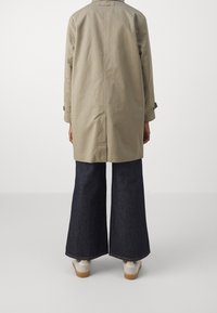 Trench-coat beige léger avec une coupe décontractée, associé à un pantalon en denim foncé évasé et des baskets blanches, vu de dos.