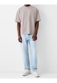 Man som bär en lös beige t-shirt, ljusblå jeans och vita sneakers som står mot en enfärgad vit bakgrund.