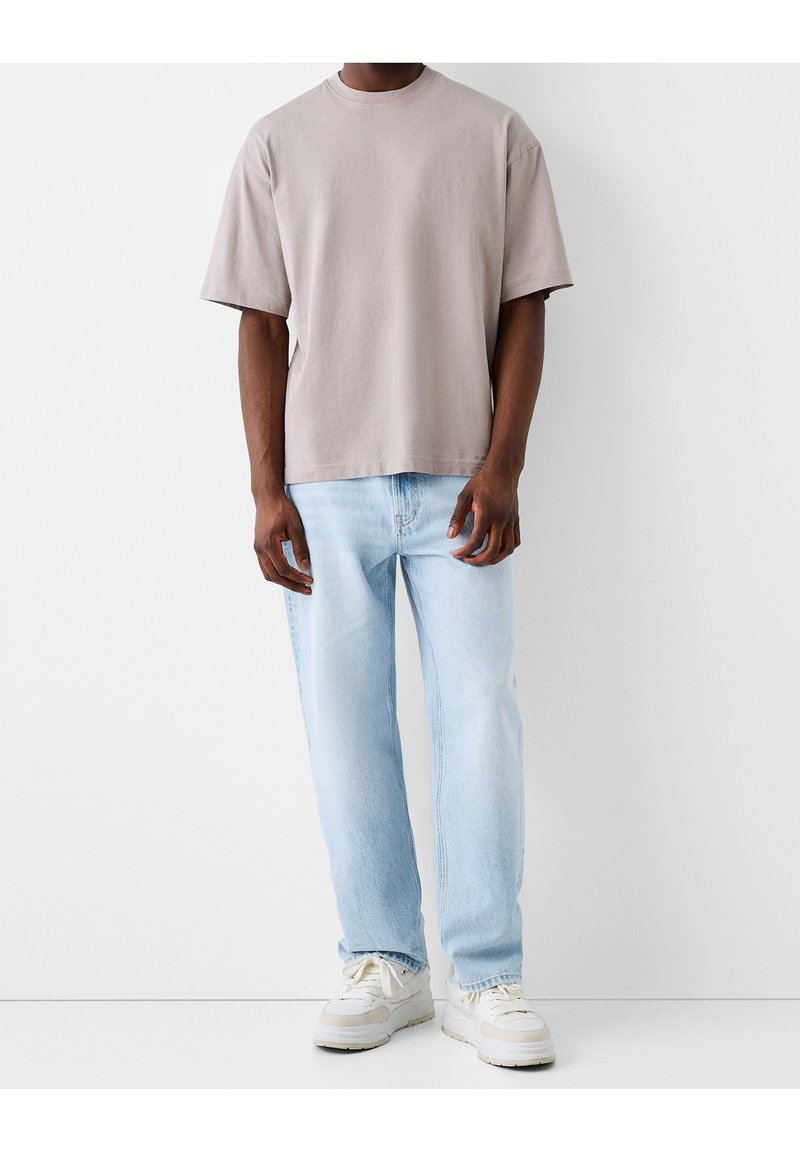 Man som bär en lös beige t-shirt, ljusblå jeans och vita sneakers som står mot en enfärgad vit bakgrund.