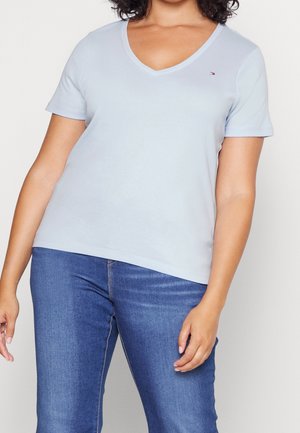 Kvinde iført lyseblå V-hals kortærmet trøje og blå jeans, stående med venstre arm afslappet og højre hånd nær låret.