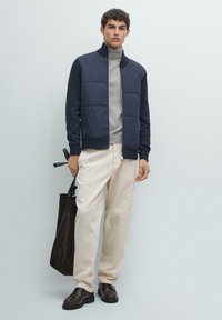 Námořnická prošívaná bunda přes šedý rolák, doplněná krémovými kalhotami a hnědými botami. Drží tmavě hnědou kabelku typu tote.