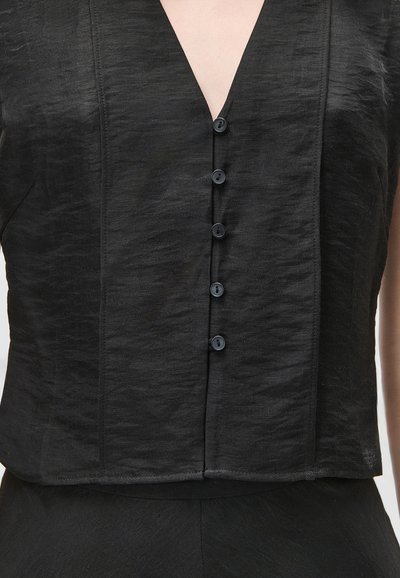 Μαύρο τοπ χωρίς μανίκια με υφή, V-λαιμόκοψη και έξι μικρά μαύρα κουμπιά που κατεβαίνουν στο μπροστινό μέρος, φορεμένο με αντίστοιχα μαύρα κάτω μέρη.