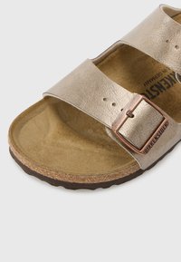 Sandalia de cuero dorado con dos correas ajustables, plantilla de ante marrón y una hebilla de metal en tono cobre. La suela de corcho añade textura.