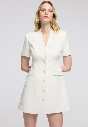 Blonde vrouw met pareloorbellen die een wit jurkje met korte mouwen, knopen, klepzakken en subtiel bloemmotief op de borst draagt.
