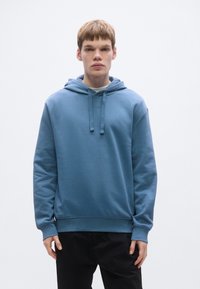 Camisola com capuz azul claro, com bolso frontal tipo canguru, capuz ajustável com cordão e punhos canelados. Tecido suave e macio. Combinada com calças pretas.