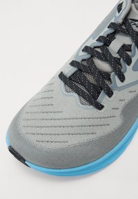 Chaussure de sport gris clair avec des accents bleus ; tige en mesh respirant, lacets noirs et surface texturée ; orteil courbé et semelle rembourrée.