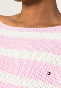 Pull en tricot aux rayures roses et blanches. Présente un design épaules dénudées, un tissu texturé et un petit logo près de l'encolure.