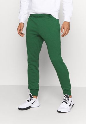 Mann trägt grüne Jogginghose, weißes Langarmshirt und weiße Nike-Sneaker und steht auf einem hellgrauen Boden.