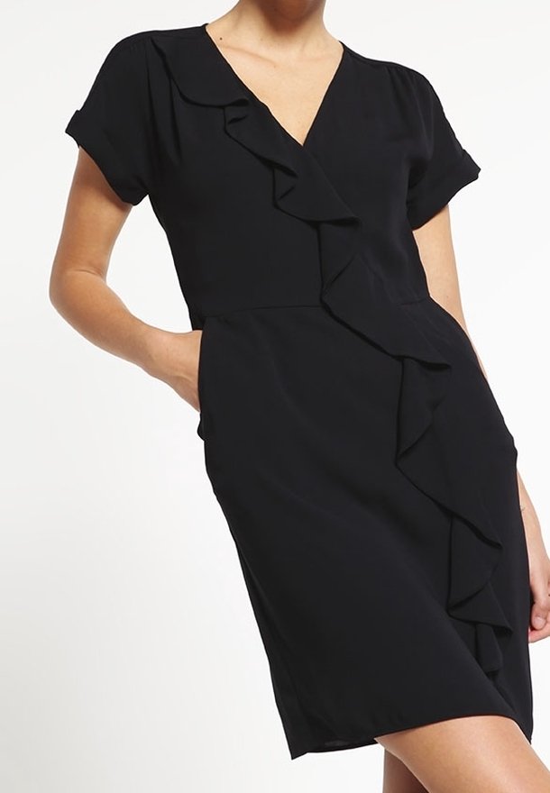 Femme portant une robe noire courte à manches courtes avec un volant sur le devant et la main dans la poche.