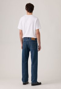 Blue denim jeans with a straight fit and visible stitching. A tan leather patch on the back waistband; worn with a plain white t-shirt.