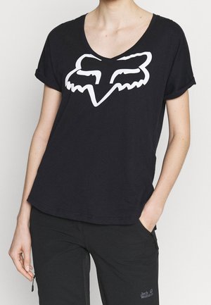 Persona vistiendo una camiseta negra de cuello en V con un logo estilizado de cabeza de zorro en blanco y pantalones negros con un pequeño logo de Jack Wolfskin.