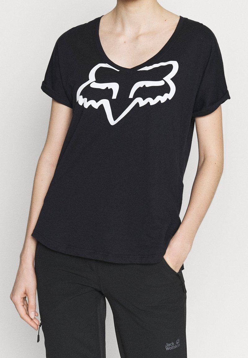 Person trägt ein schwarzes V-Ausschnitt-T-Shirt mit einem weißen, stilisierten Fuchskopf-Logo und schwarze Hosen mit einem kleinen Jack-Wolfskin-Logo.