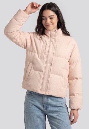 Frau mit langen dunklen Haaren, die eine hellrosa gesteppte Daunenjacke und hellblaue Jeans trägt und ihren rechten Arm leicht hebt.
