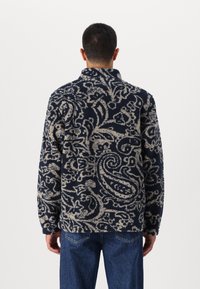 Tengerés kék polárdzseki krém színű paisley mintával, magas gallérral és hosszú ujjakkal. Puha tapintás, laza szabás, látható záródások nélkül.