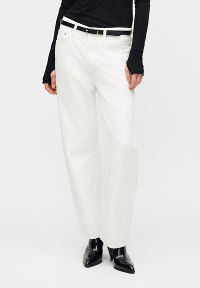 Άτομο που φορά ψηλόμεσα λευκά φαρδιά τζιν, μαύρη ζώνη, μαύρο μακρυμάνικο τοπ και μαύρα μυτερά μπότες με τακούνι.