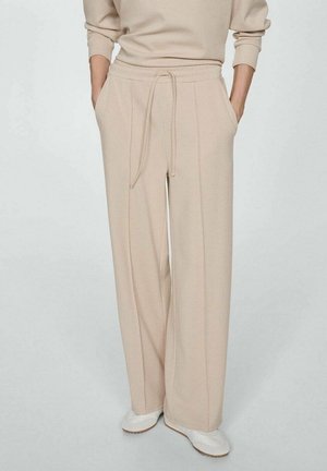 Beige Hosen mit weitem Bein, vorn mit Nähten und einem Kordelzug in der Taille, kombiniert mit einem passenden beigen Oberteil und weißen Turnschuhen, die Hände in den Taschen.