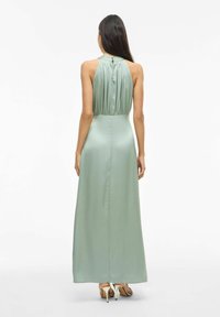 Długa, zielona satynowa sukienka z plisowanym tyłem, wysokim dekoltem i zapięciem na guziki z kluczem. Gładka faktura i płynny krój.