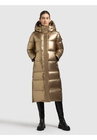 Frau trägt einen langen, glänzenden bronzefarbenen Steppmantel mit Kapuze und Händen in den Taschen, kombiniert mit schwarzen kniehohen Stiefeln vor einfachem Hintergrund.