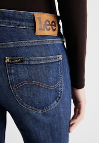 Tmavomodré džínsy z denimu s koženou nášivkou Lee na páse, so žltým stehom a zadnou vreckom s zaoblenými švami.