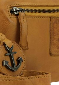 Primo piano di una borsa in pelle color sabbia con un'emblema ad ancora in metallo e una tasca con zip cucita, dotata di una linguetta in pelle.