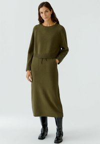 Olivengrünes Strickkleid mit langen Ärmeln, geripptem Ausschnitt und Taille, zwei vorderen Taschen und geradem Schnitt, kombiniert mit schwarzen, glänzenden Stiefeln.
