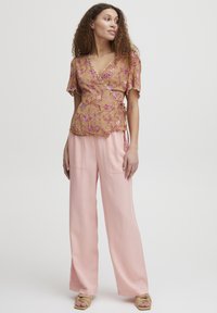 Blusa a maniche corte avvolgente con ricami floreali in oro e rosa, abbinata a pantaloni rosa a gamba larga e tacchi beige.