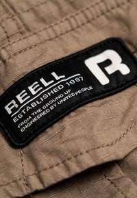 Beiger Stoff mit schwarzem rechteckigem Aufnäher mit der Aufschrift „REELL Established 1997 From the ground up Engineered by united people“ und einem weißen „R“-Logo.