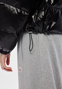 Melns pūkains mētelis ar spīdīgu tekstūru, elastīgiem piedurkņu galiem, kontrastējošas pelēkas bikses, neliels logotipa uzlīme un regulējams šnīkeris jostasvietā.