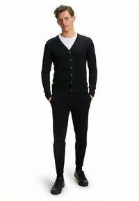 Cardigan nero con scollo a V, chiusura frontale con bottoni e design aderente, abbinato a pantaloni neri slim fit e sneakers nere. Tessuto uniforme e liscio su tutta la superficie.