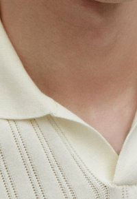 Gros plan sur le cou et le haut de la poitrine d'une personne portant un pull en tricot crème avec un col rabattu.