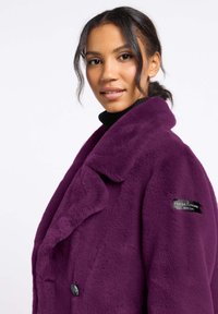Fioletowy futrzany płaszcz z wyprofilowanym kołnierzem, podwójnymi guzikami i czarną etykietą z tkaniny na rękawie. Tekstura i miękki wygląd.