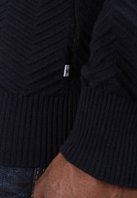 Marineblå strikket genser med et teksturert herringbone-mønster, ribbestrikkede mansjetter og en liten merkeetikett på siden.