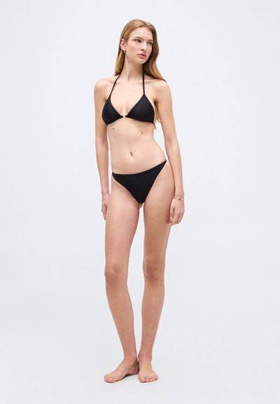 Γυναίκα με μακριά μαλλιά που φοράει μαύρο μαγιό τύπου halter, όρθια ξυπόλητη μπροστά σε απλό γκρι ανοιχτό φόντο, κοιτάζοντας προς τα πλάγια.