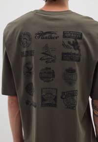 Camiseta de manga corta en verde oliva con diversos gráficos impresos en negro, incluyendo logotipos e ilustraciones, en la parte trasera.