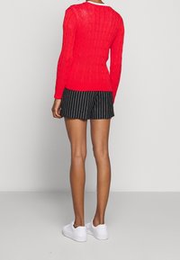 Röd stickad tröja med flätmönster, ribbade muddar och midja, i kombination med svarta randiga shorts. Modellen bär vita sneakers.